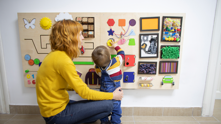 En kvinna sitter på huk framför en anslagstavla på en vägg. Barnet framför henne leker med de uppsatta leksakerna på tavlan.