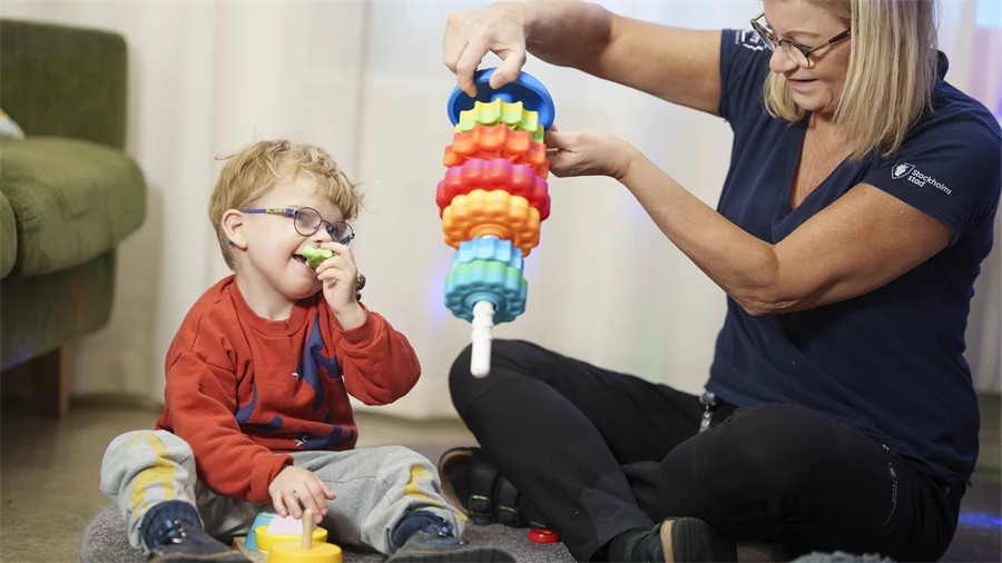 En pedagog sitter på golvet och leker med ett barn. Båda ler och tittar på en leksak.