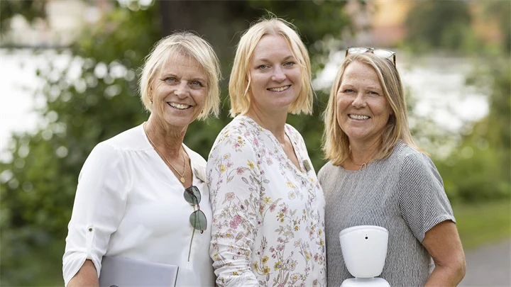 Tre kvinnor står utomhus. De tittar in i kameran och ler. En kvinna håller i ett lärverktyg.