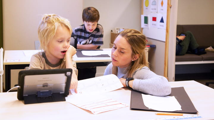 En lärare sitter bredvid en elev som arbetar med en surfplatta och pratar om någon uppgift. Bakom dem sitter an annan elev och läser.