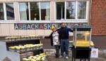 På bilden ser du två personer som håller om varandra framför en skylt där det står Åsbackaskolan i olika färger. Bredvid dem står en popcornmaskin.
