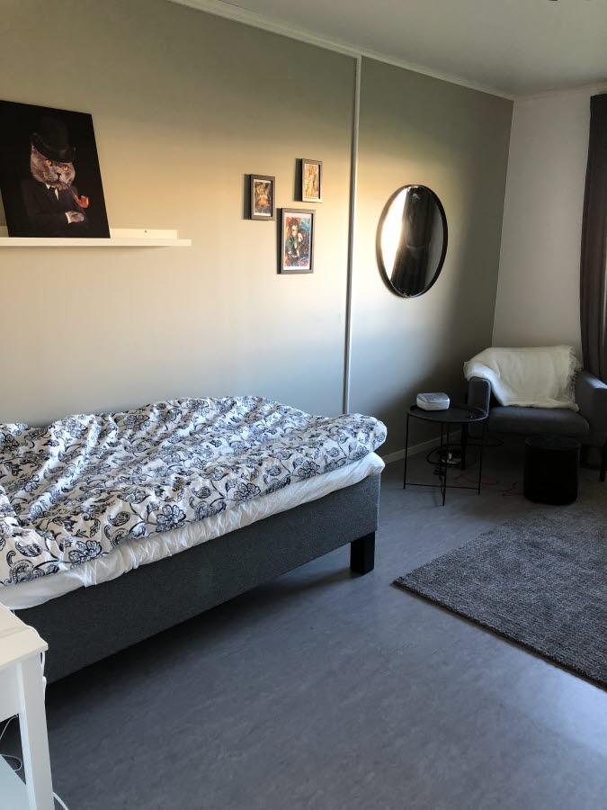 En bäddad säng med några tavlor och en spegel på väggen samt en fåtölj vid hörnet.