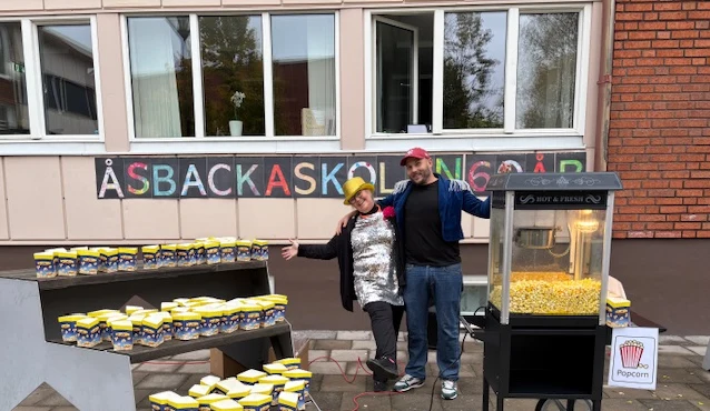 På bilden ser du två personer som håller om varandra framför en skylt där det står Åsbackaskolan i olika färger. Bredvid dem står en popcornmaskin.